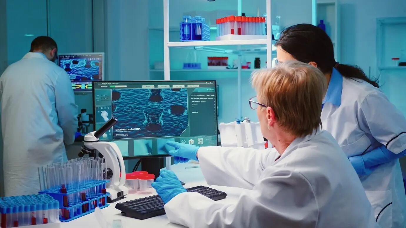 Старший биохимик в современной оснащённой лаборатории записывает на компьютере изменения в составе вакцины, консультируясь с коллегой.