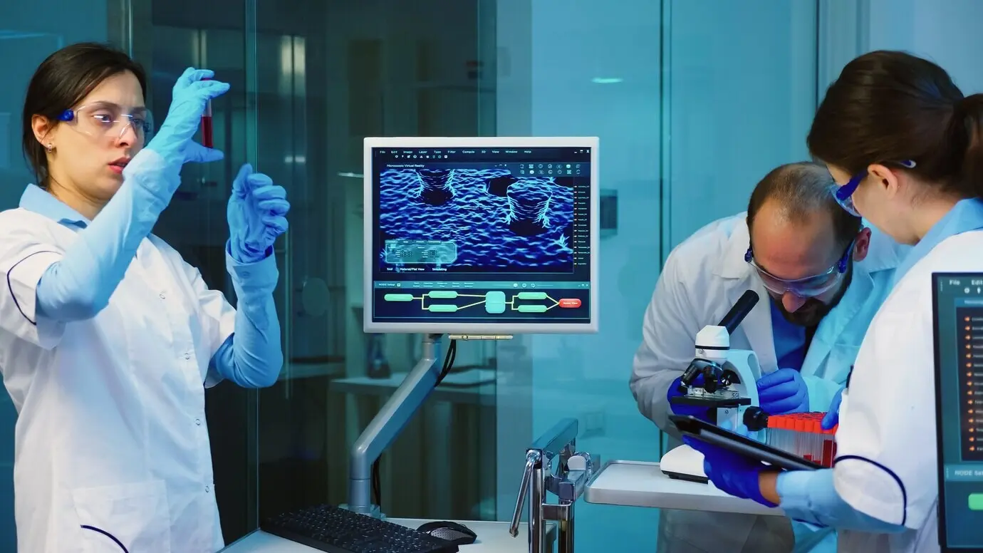 Старший химик проводит анализ образца крови, пока коллеги обсуждают.